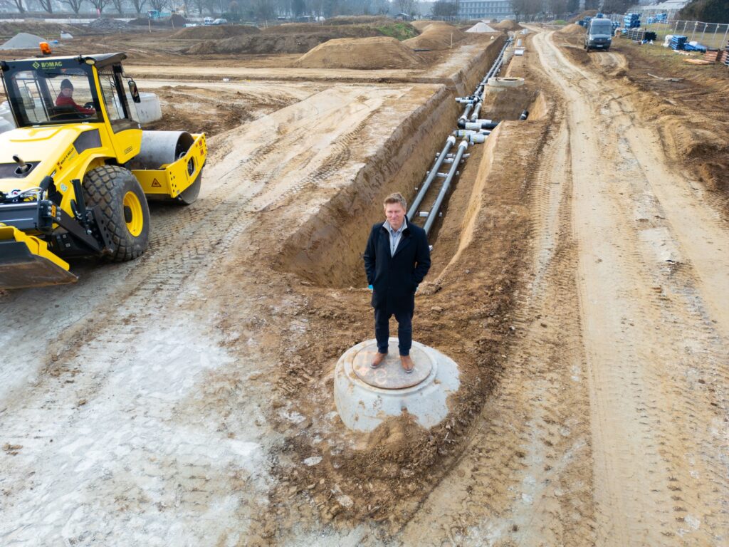 Mann steht auf einer Baustelle mit Fernwärmerohren und links ist ein Bagger zu sehen
